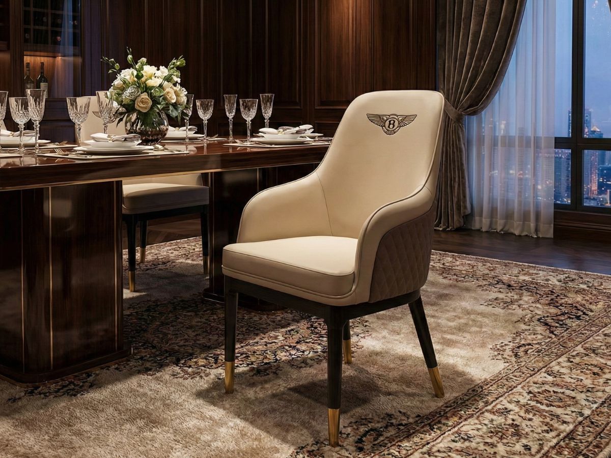 Ghế ăn Bentley bọc da cao cấp màu kem nhạt, đặt nổi bật trước bàn ăn gỗ sẫm màu trong phòng ăn luxury. Lưng ghế thêu logo…