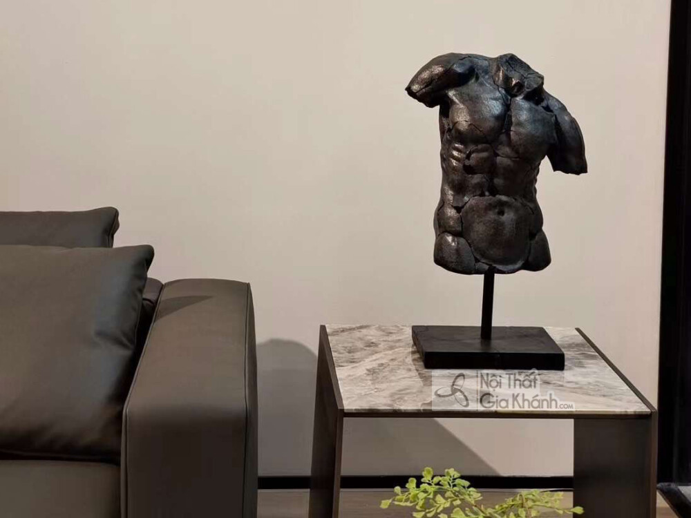 Cận cảnh bàn phụ đặt cạnh sofa, có mặt bàn bằng đá marble vân xám trắng và chân đế màu đen. Trên bàn đặt một bức tượng bán…