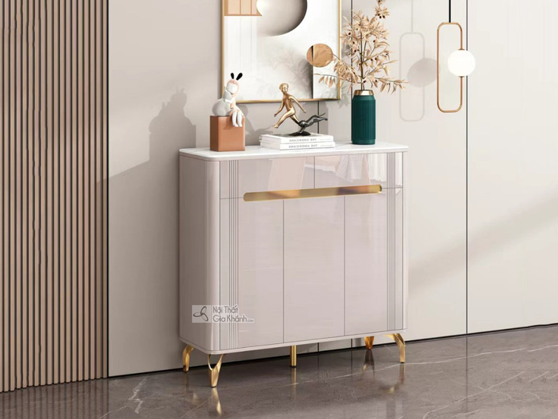 Tủ giày hiện đại TDEH165 màu kem bóng, đặt ở góc tiền sảnh. Tủ có mặt trên bằng đá màu trắng, ba cánh cửa tủ với các đường…