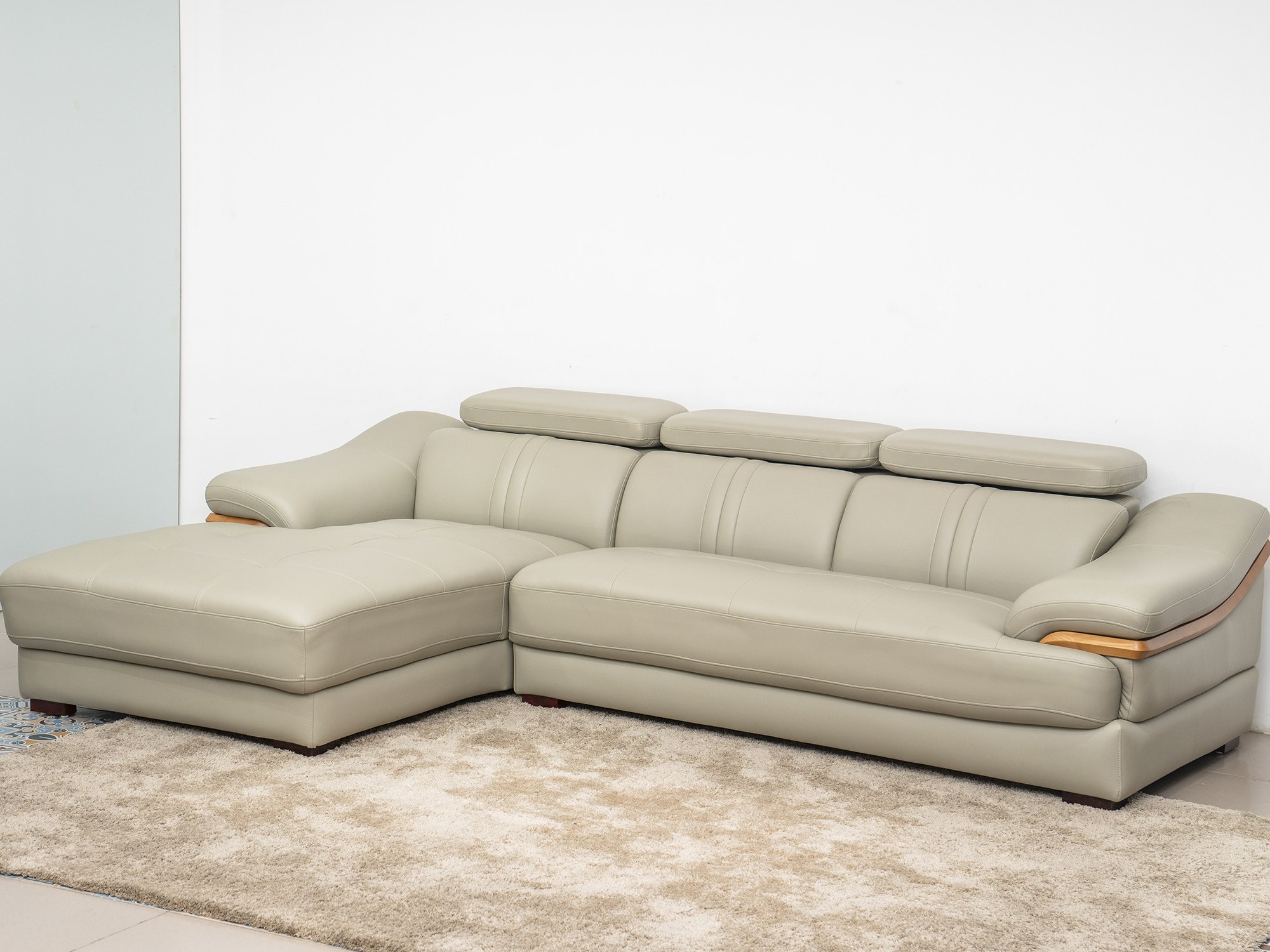 Sofa da góc phải màu be/ghi sáng, chụp từ góc cao phía trước bên trái, đặt trên thảm lông màu kem và sàn gạch. Tựa đầu đượ…
