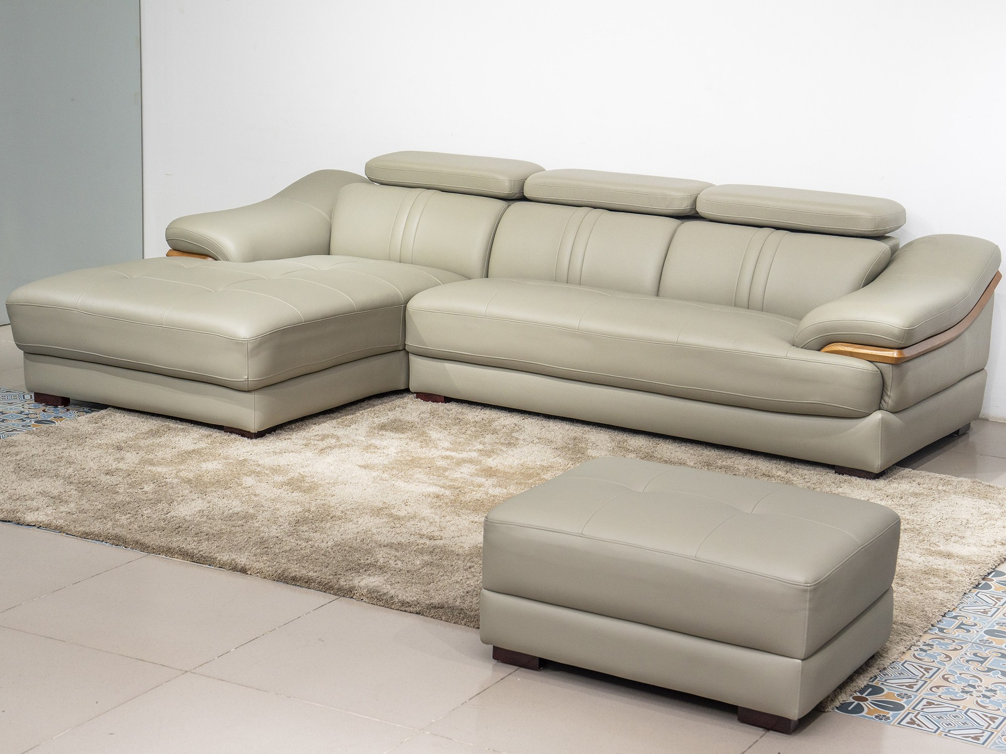 Sofa da góc phải màu be/ghi sáng, chụp từ góc cao phía trước bên trái, đặt trên thảm lông màu kem và sàn gạch. Kèm theo mộ…