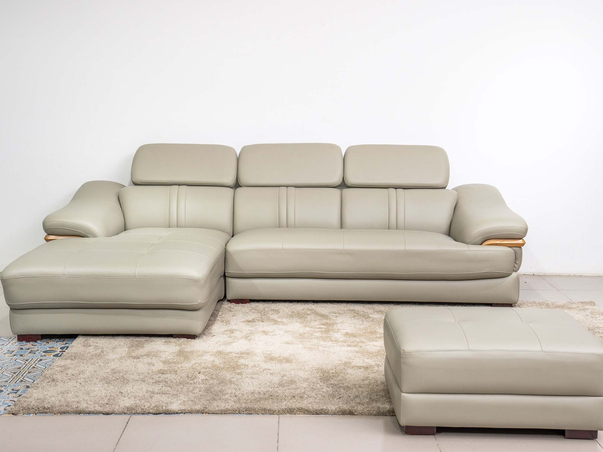 Sofa da góc phải màu be/ghi sáng, chụp từ góc thấp phía trước, đặt trên thảm lông màu kem và sàn gạch. Kèm theo một đôn nh…