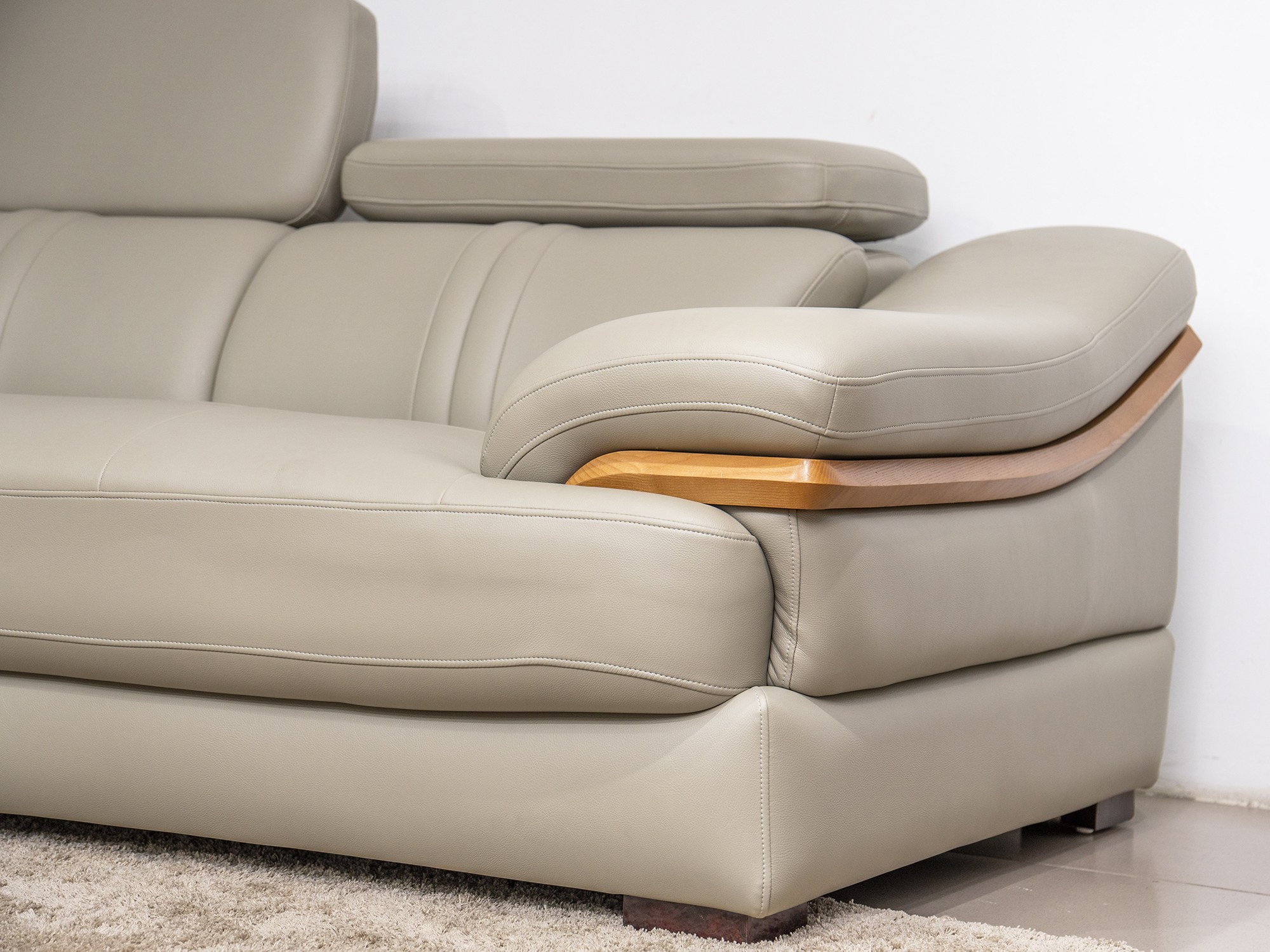 Cận cảnh tựa tay bo cong của sofa da màu be/ghi sáng, với đường may nổi bật và chi tiết ốp gỗ màu nâu sáng. Chụp từ góc ng…