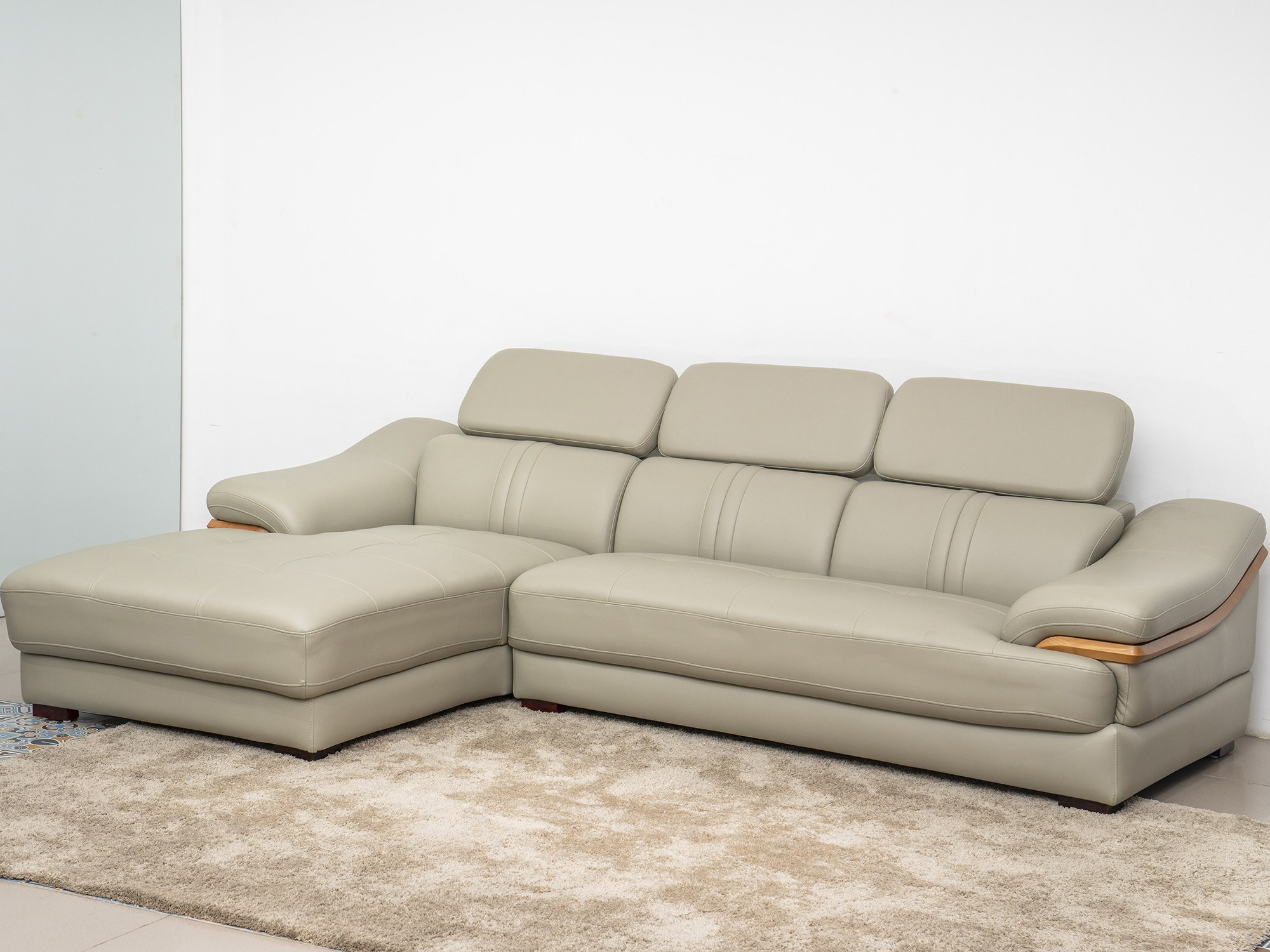 Sofa da góc phải màu be/ghi sáng, chụp từ góc thấp phía trước bên trái, đặt trên thảm lông màu kem trong không gian phòng…
