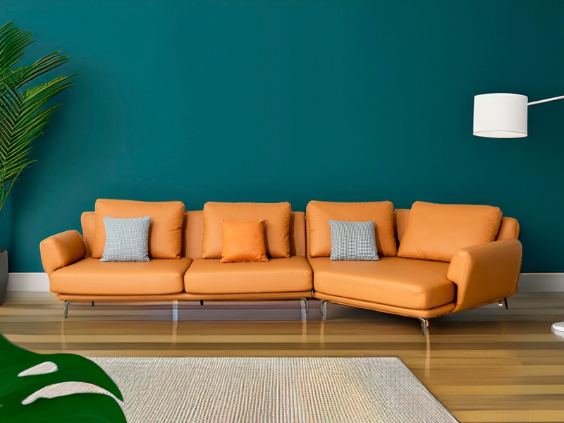Bộ sofa da góc màu cam được chụp chính diện, đặt trên sàn gỗ sáng màu với thảm trải sàn họa tiết, nổi bật trên nền tường x…