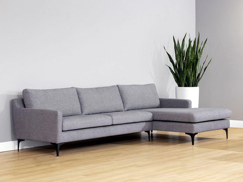 Sofa lệch góc bọc vải cao cấp màu xám đậm, thiết kế thanh mảnh và hiện đại. Sản phẩm nổi bật trên nền tường xám nhạt và sà…