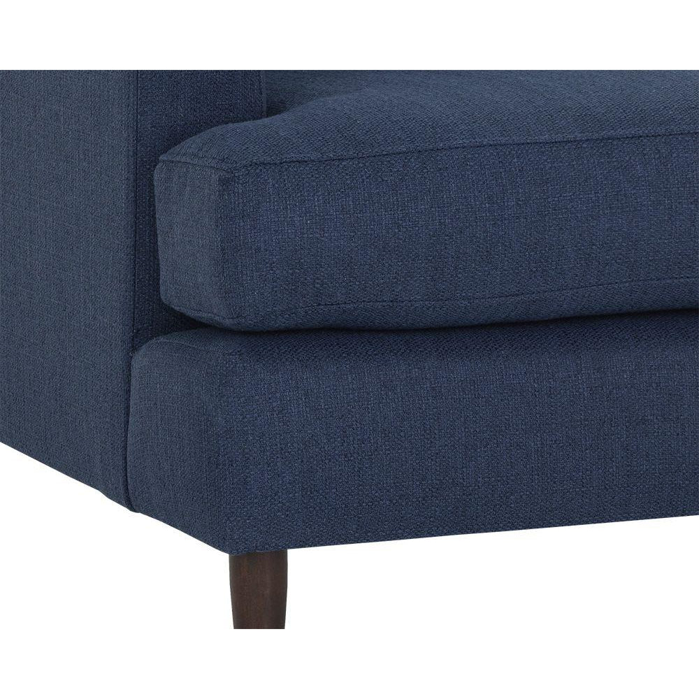 Cận cảnh phần chân ghế gỗ walnut màu nâu sẫm và góc đệm ngồi của sofa băng bọc vải màu xanh đậm. Hình ảnh làm nổi bật chất…
