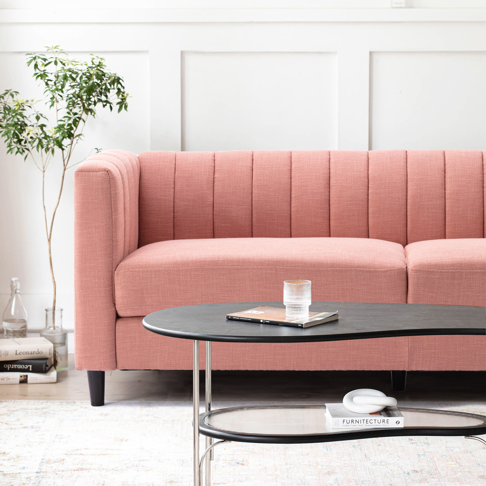 Sofa băng bọc vải màu hồng nhạt được chụp từ góc nghiêng, làm nổi bật thiết kế tựa lưng có rãnh dọc và tay vịn cao. Phía t…