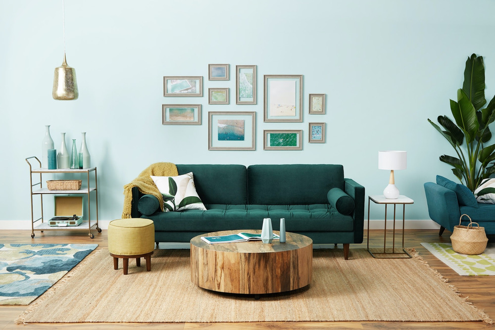 Sofa băng 3 chỗ bọc vải màu xanh lá cây đậm, đặt trong phòng khách ấm cúng với tường xanh nhạt, sàn gỗ và thảm họa tiết. G…