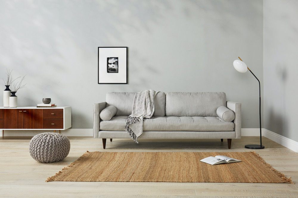 Sofa băng 3 chỗ bọc vải màu xám nhạt, đặt trong phòng khách tối giản với tường trắng, sàn gỗ và thảm cói. Góc chụp ngang…
