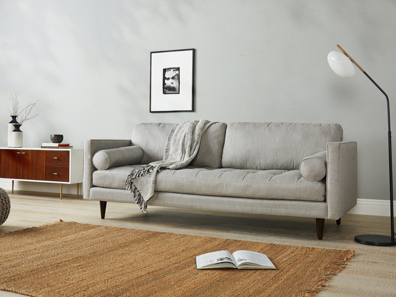 Sofa băng 3 chỗ bọc vải màu xám nhạt, đặt trong phòng khách hiện đại với tường trắng, sàn gỗ và thảm cói. Góc chụp ngang…