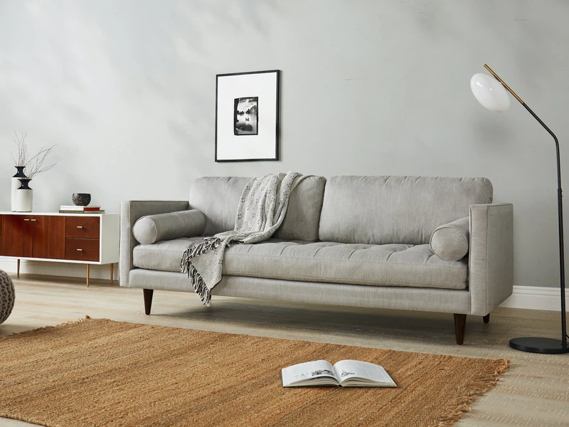 Sofa băng 3 chỗ bọc vải màu xám nhạt, đặt trong phòng khách hiện đại với tường trắng, sàn gỗ và thảm cói. Góc chụp ngang…