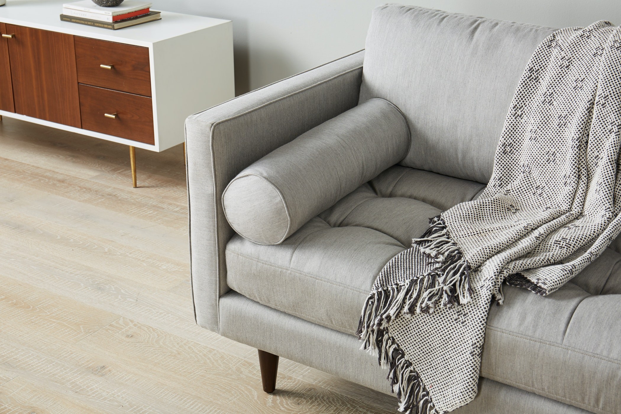 Cận cảnh tay vịn và gối ôm tròn của sofa bọc vải màu xám nhạt, đặt trong phòng khách hiện đại. Chi tiết đường may tinh tế…