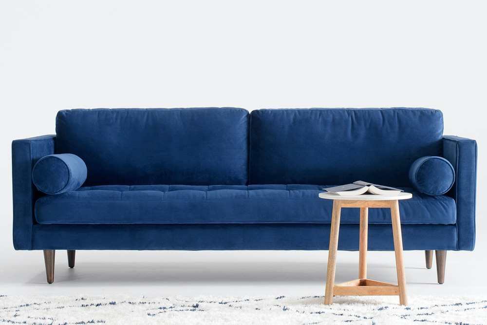 Sofa băng 3 chỗ bọc vải màu xanh navy nổi bật, đặt giữa phòng khách với thảm họa tiết và bàn trà gỗ nhỏ. Góc chụp chính di…