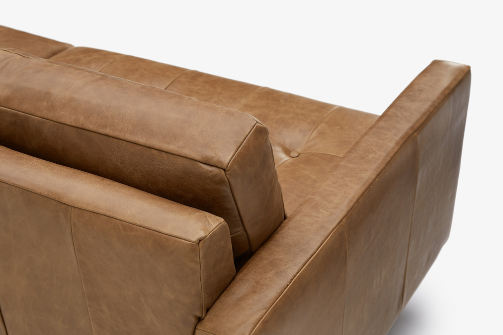 Cận cảnh góc tựa lưng và tay vịn của sofa băng bọc da màu nâu caramel. Hình ảnh tập trung vào đường may tinh xảo, chất liệ…