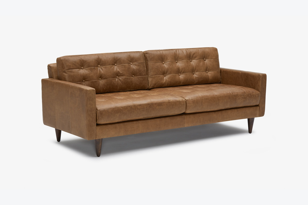 Sofa băng bọc da màu nâu caramel, chụp từ góc thấp phía trước trên nền trắng. Hình ảnh làm nổi bật dáng vẻ thanh lịch và h…