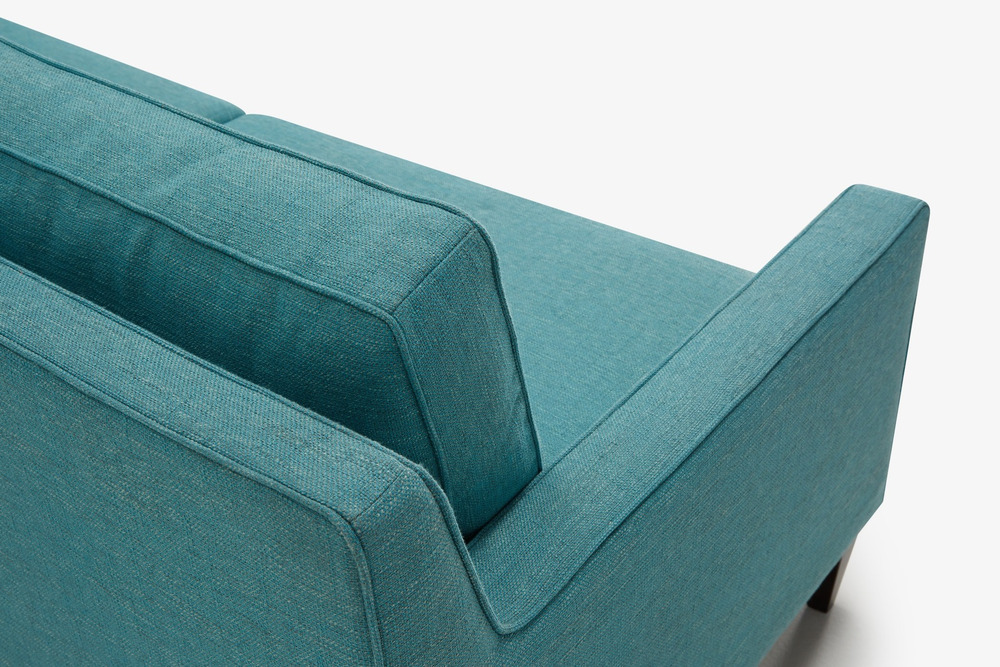 Cận cảnh phần tay vịn và tựa lưng của sofa băng bọc vải màu xanh ngọc, chụp từ góc cao phía sau. Chất liệu vải dệt mịn với…