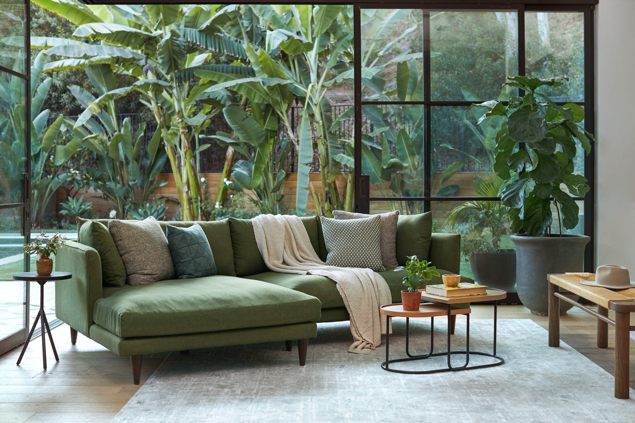 Sofa góc chữ L Lewis SF-489 bọc vải màu xanh lá cây đậm, đặt trong phòng khách có cửa kính lớn nhìn ra khu vườn nhiệt đới…