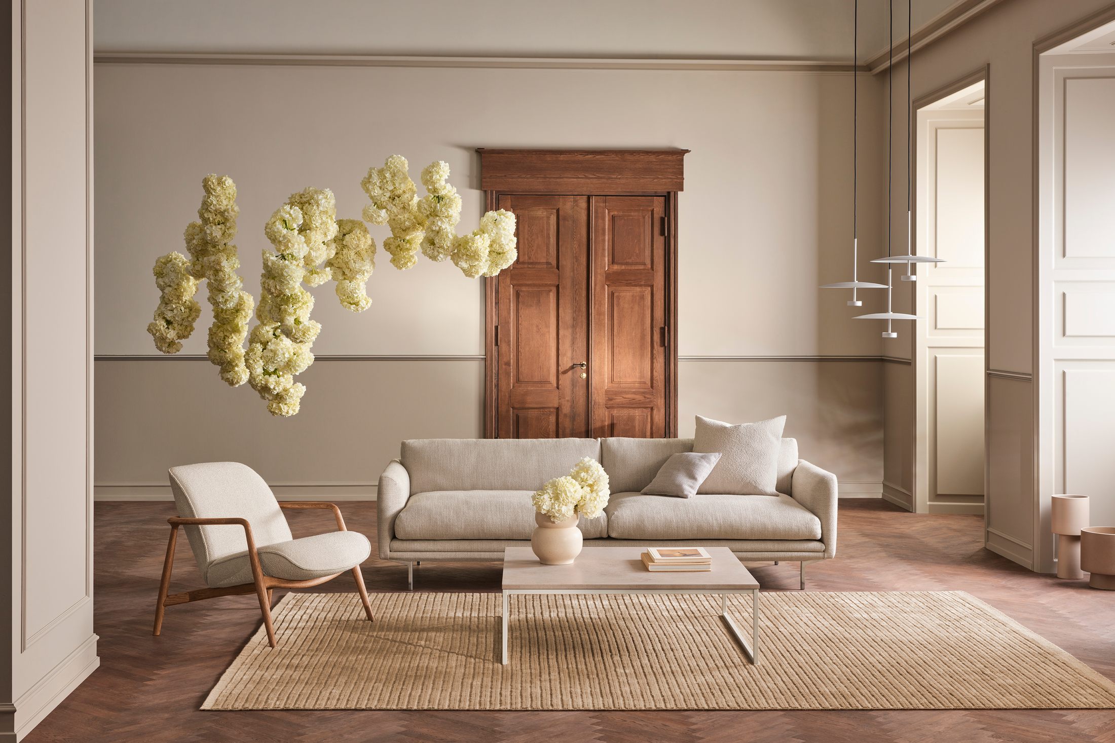 Sofa bọc vải màu be nhạt nổi bật trong không gian phòng khách tân cổ điển, với tường màu kem và sàn gỗ xương cá. Bố cục ba…