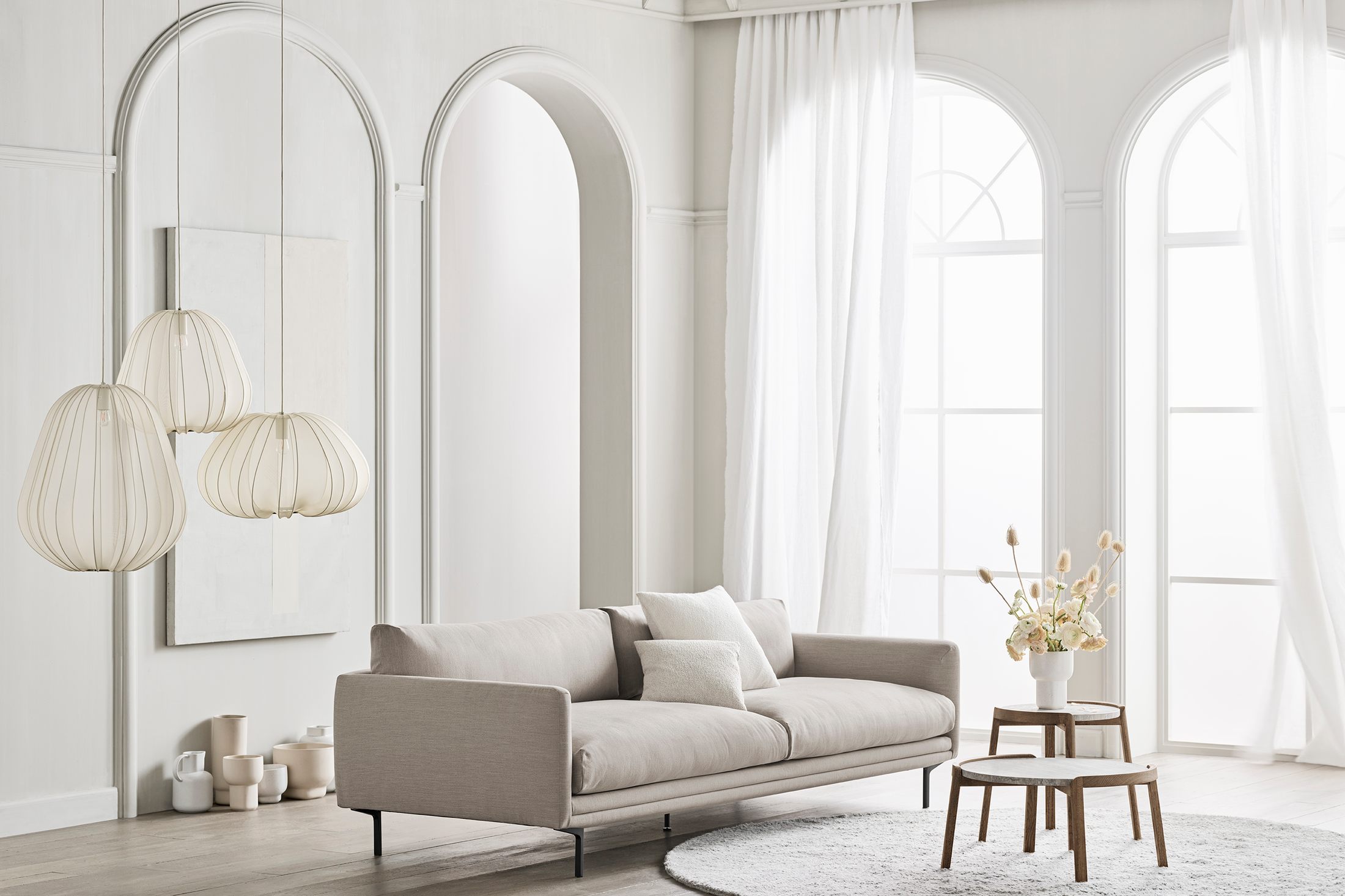 Sofa băng bọc vải màu be nhạt, kiểu dáng đương đại, đặt trong không gian phòng khách hiện đại với tường trắng, cửa vòm lớn…