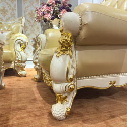 Cận cảnh chân ghế sofa với các đường cong uốn lượn và họa tiết chạm khắc dát vàng tinh xảo trên nền trắng ngà. Phần đệm ng…