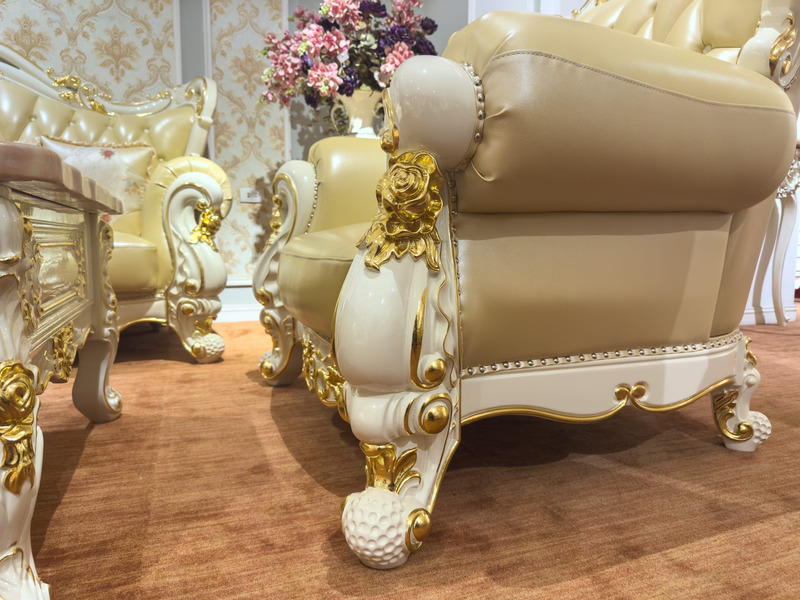 Cận cảnh chân ghế sofa với các đường cong uốn lượn và họa tiết chạm khắc mạ vàng tinh xảo trên nền trắng ngà. Phần đệm ng…