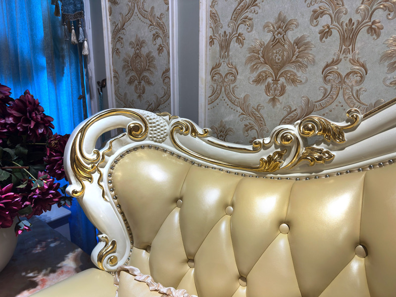 Chi tiết phần lưng tựa và tay vịn của sofa, với các đường cong mềm mại và họa tiết mạ vàng nổi bật trên nền da màu kem. N…
