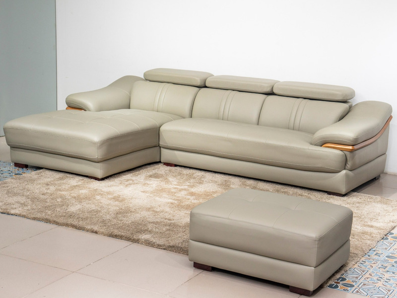 Sofa da góc phải màu be/ghi sáng, chụp từ góc cao phía trước bên phải, đặt trên thảm lông màu kem và sàn gạch. Kèm theo mộ…