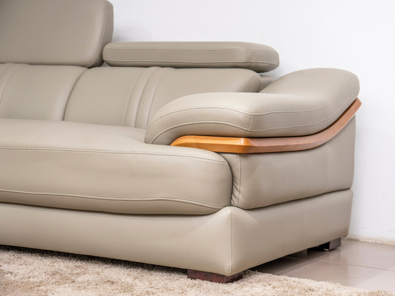 Cận cảnh tựa tay bo cong của sofa da màu be/ghi sáng, có chi tiết ốp gỗ màu nâu sáng. Chụp từ góc thấp, làm nổi bật đường…