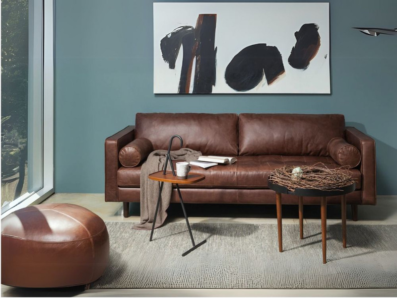 Phòng khách hiện đại với sofa băng màu nâu đậm, bàn trà gỗ tròn, đôn da và tranh trừu tượng trên tường màu xanh. Không gia…