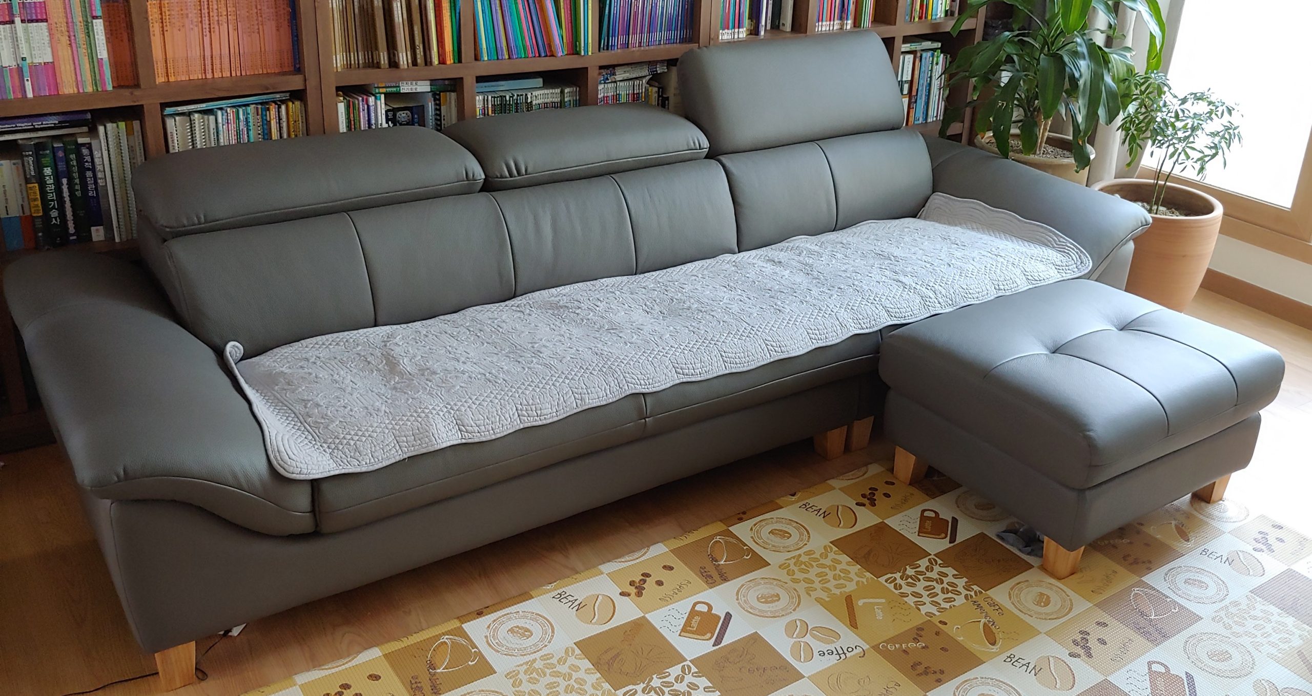 Sofa Haven màu xám nhạt, cấu hình chữ L kết hợp đôn rời, đặt trong không gian phòng khách hiện đại với kệ sách lớn phía sa…