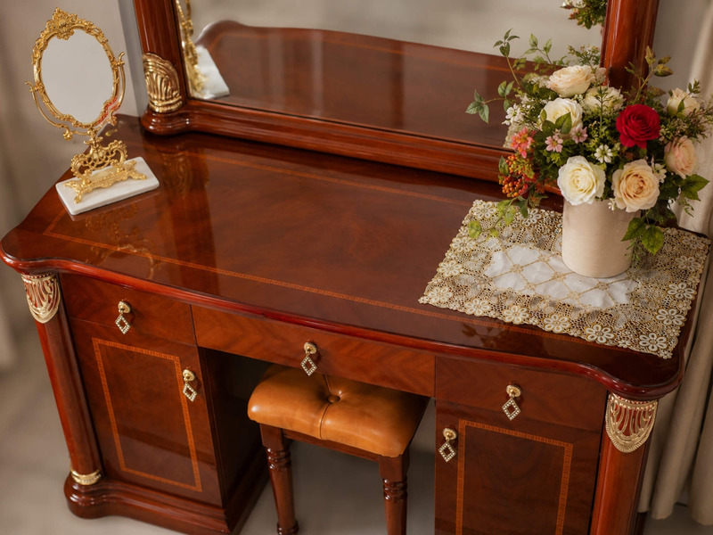Góc nhìn cận cảnh mặt bàn trang điểm màu nâu đỏ, bề mặt bóng loáng phản chiếu ánh sáng. Bàn có hai ngăn kéo nhỏ và một ngă…