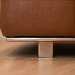 Cận cảnh chân sofa làm từ inox sáng bóng, thiết kế hình chữ nhật vững chắc, nâng đỡ phần thân sofa màu nâu da bò. Chi tiết…