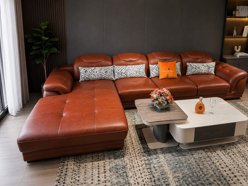 Sofa góc chữ L màu nâu da bò nhìn từ phía trước, tập trung vào phần ghế dài thư giãn và các gối tựa họa tiết. Sản phẩm đượ…