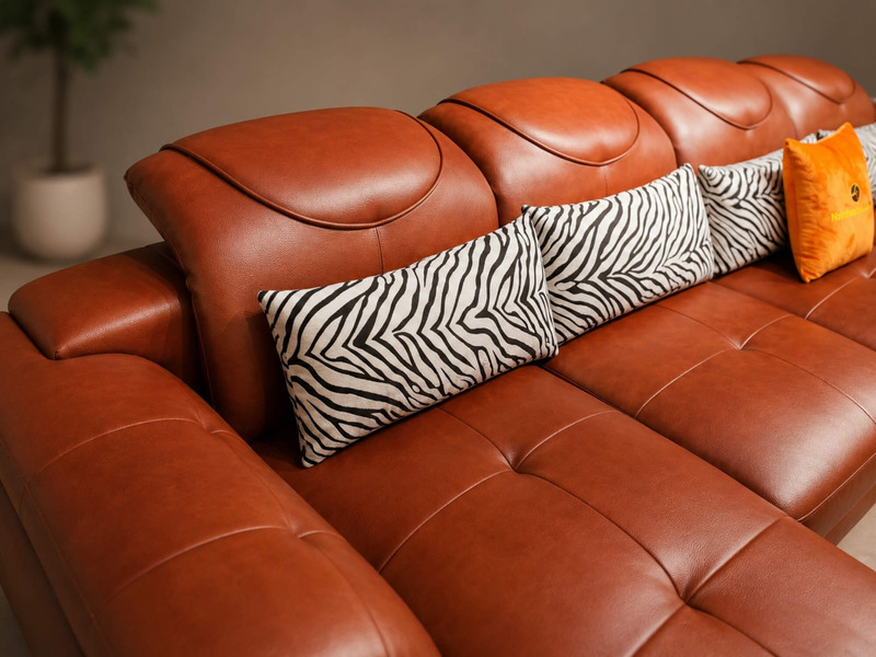 Cận cảnh phần lưng và tay vịn của sofa góc màu nâu da bò, với các đường may tạo điểm nhấn và gối tựa họa tiết ngựa vằn. Th…